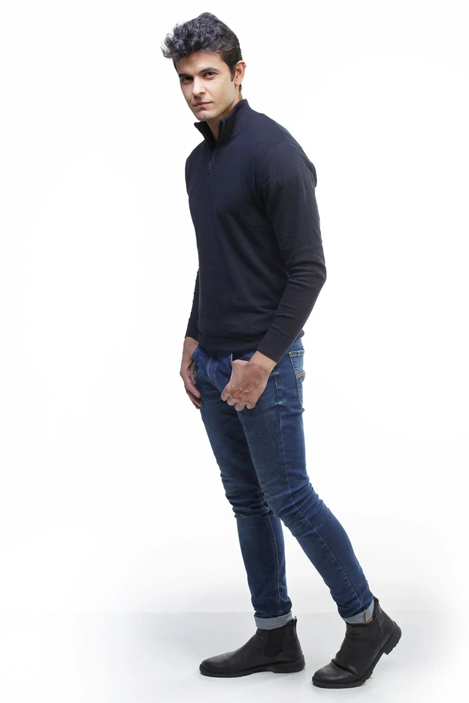 Hombre con suéter tejido azul marino, jeans ajustados y botines negros, fondo blanco.